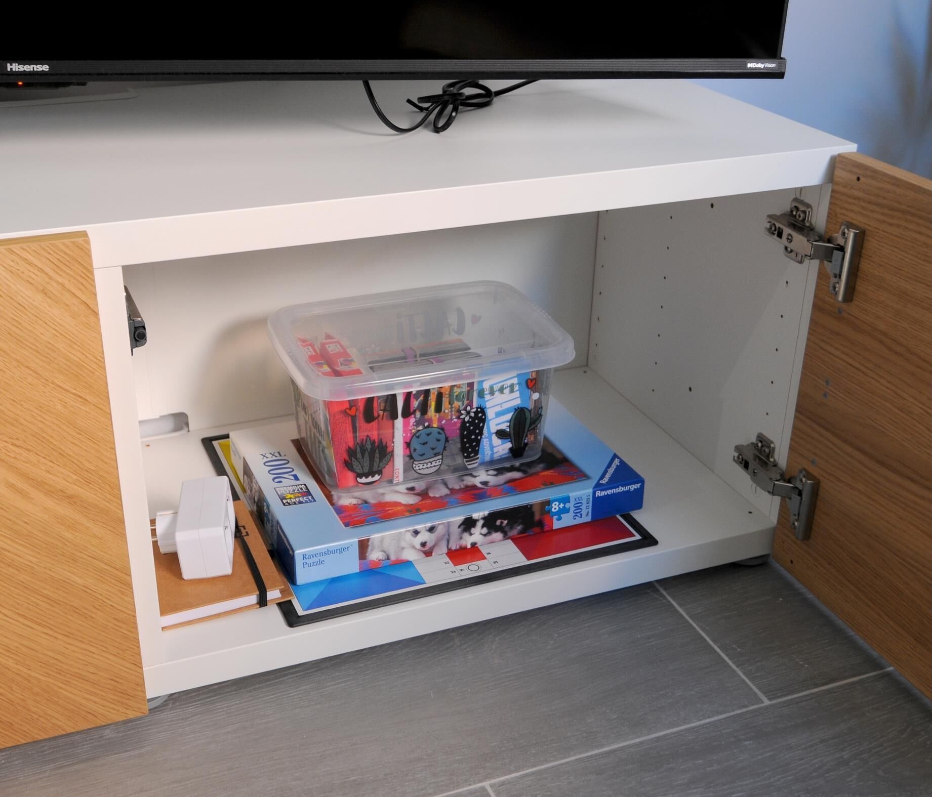 Een witte kast met een flatscreen televisie erboven bevestigd. Binnenin de kast bevindt zich een transparante plastic opbergdoos met een puzzelspel en een wit voorwerp.