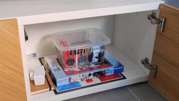 Een witte kast met een flatscreen televisie erboven bevestigd. Binnenin de kast bevindt zich een transparante plastic opbergdoos met een puzzelspel en een wit voorwerp.
