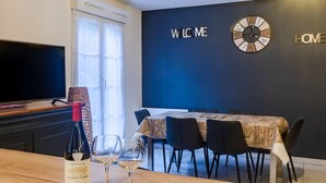 Un coin repas moderne avec une table en bois, deux verres à vin et une bouteille de vin. Une télévision se trouve à gauche, et une horloge murale est placée au-dessus de la table. Le mur porte une inscription « BIENVENUE » et une autre « MAISON ».