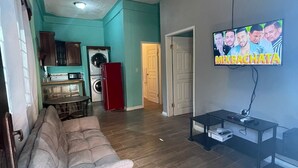 Una sala de estar con un sofá, un ventilador de techo, un televisor y una zona de cocina al fondo.