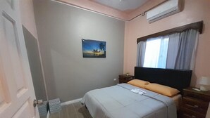 Un dormitorio con cama, mesita de noche, ventana con cortinas, aire acondicionado de pared y un cuadro en la pared.