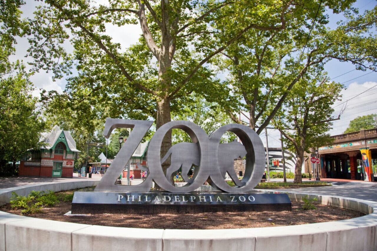 A large metallic sign with the word "ZOO" and "Philadelphia Zoo" inscribed on it, surrounded by a landscaped area with trees.