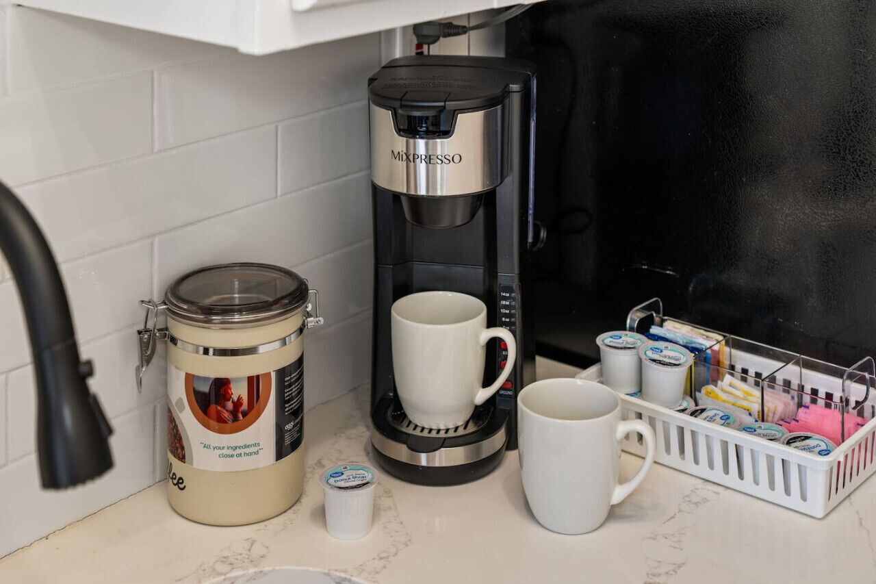 A coffee maker, a jar of coffee, a mug, and a basket of coffee pods on a kitchen counter.