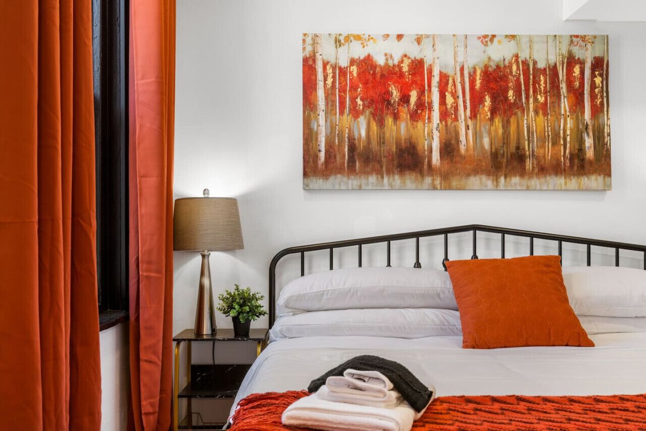 A neatly made bed with white linens and an orange pillow, a painting of a forest on the wall, and a lamp on a bedside table.