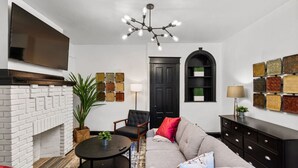 A modern living room with a white brick fireplace, a black TV mounted on the wall, a grey sofa, a black armchair, and a round coffee table. - Modern 3BR Philadelphia Stay, Central Location & Free Parking (Philadelphia)