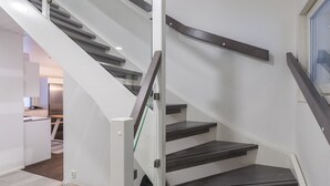 Una escalera moderna con barandilla de vidrio y una cocina al fondo.