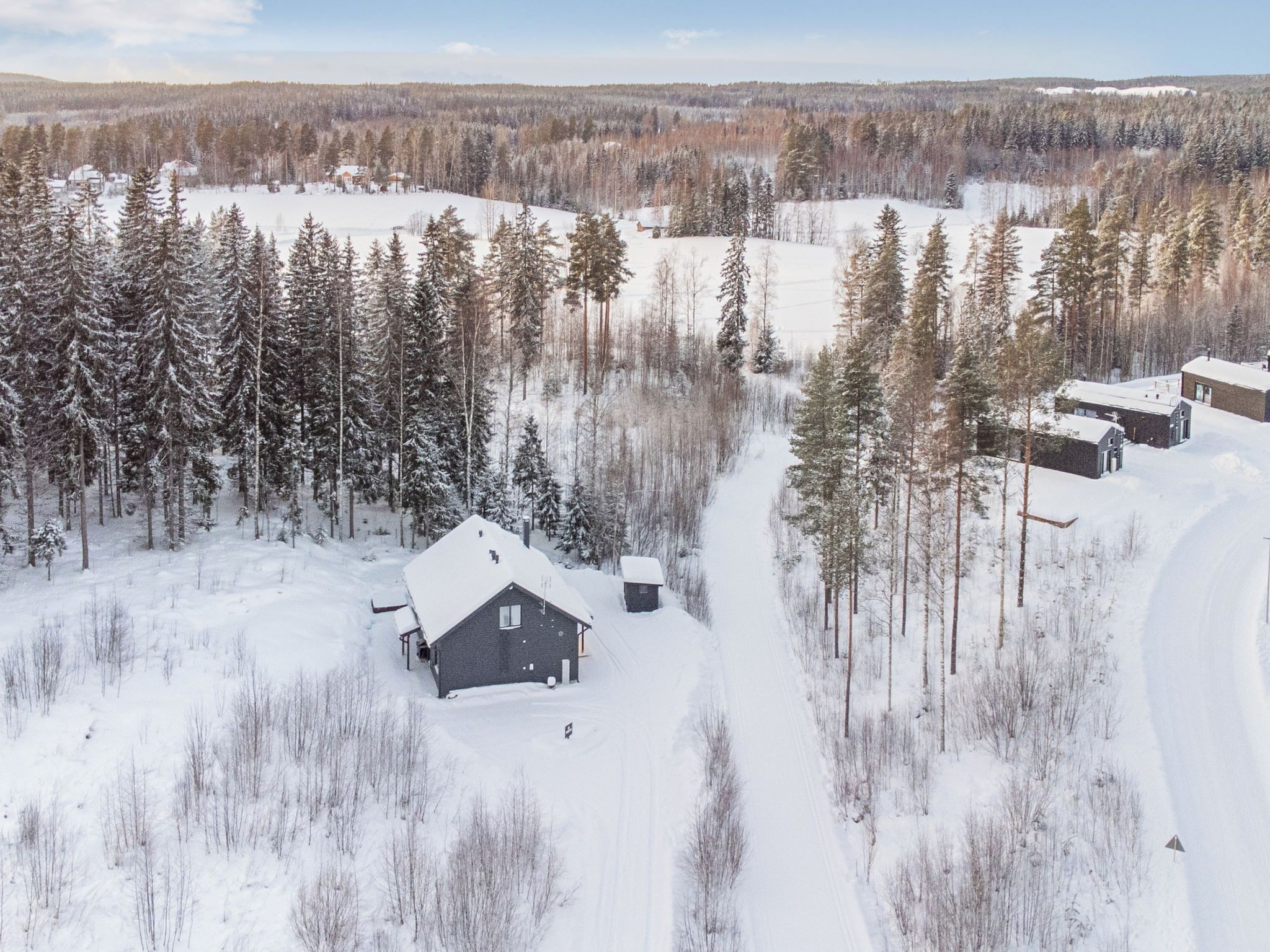 Un paysage enneigé avec une cabane et plusieurs bâtiments plus petits, le tout entouré de forêts denses.