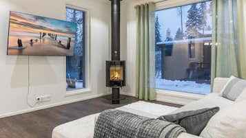 Ein Wohnzimmer mit Kamin, einem an der Wand montierten Fernseher und großen, mit Vorhängen versehenen Fenstern.