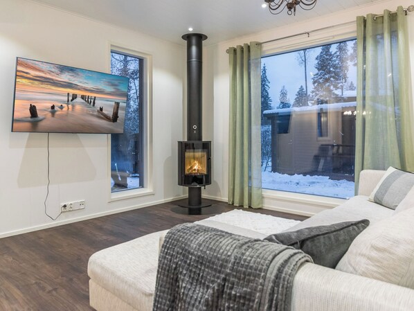 Una sala de estar con chimenea, un televisor montado en la pared y grandes ventanales con cortinas.