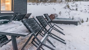 Un área de estar al aire libre con una mesa y sillas cubiertas de nieve, un jacuzzi y un edificio con una ventana iluminada al fondo.