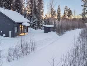 Una cabaña con techo nevado, un pequeño porche y un sendero despejado que conduce a una estructura de madera en un bosque cubierto de nieve.