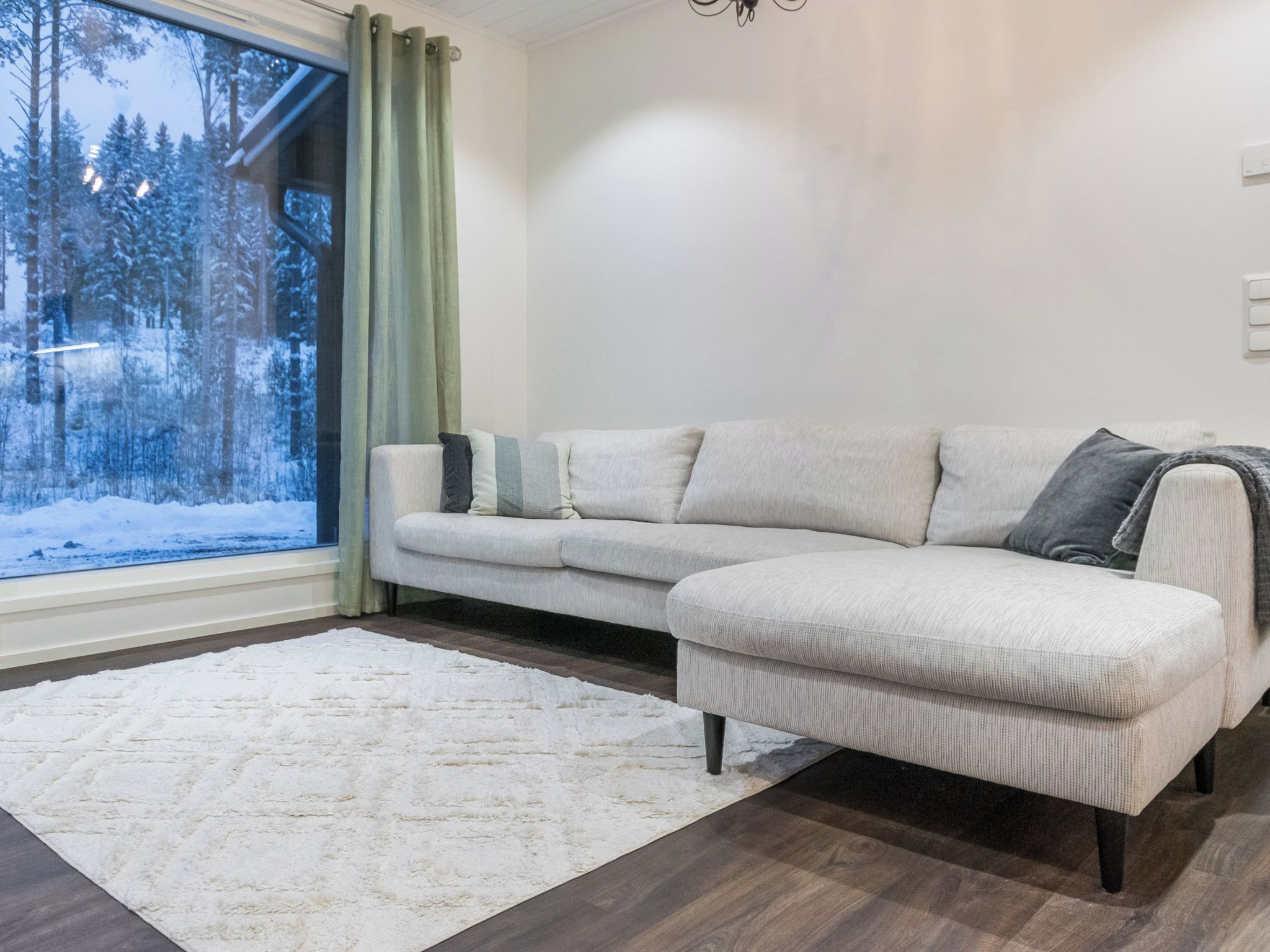 Un salon moderne avec un canapé d’angle, un tapis blanc et une vue sur une forêt enneigée par la fenêtre.