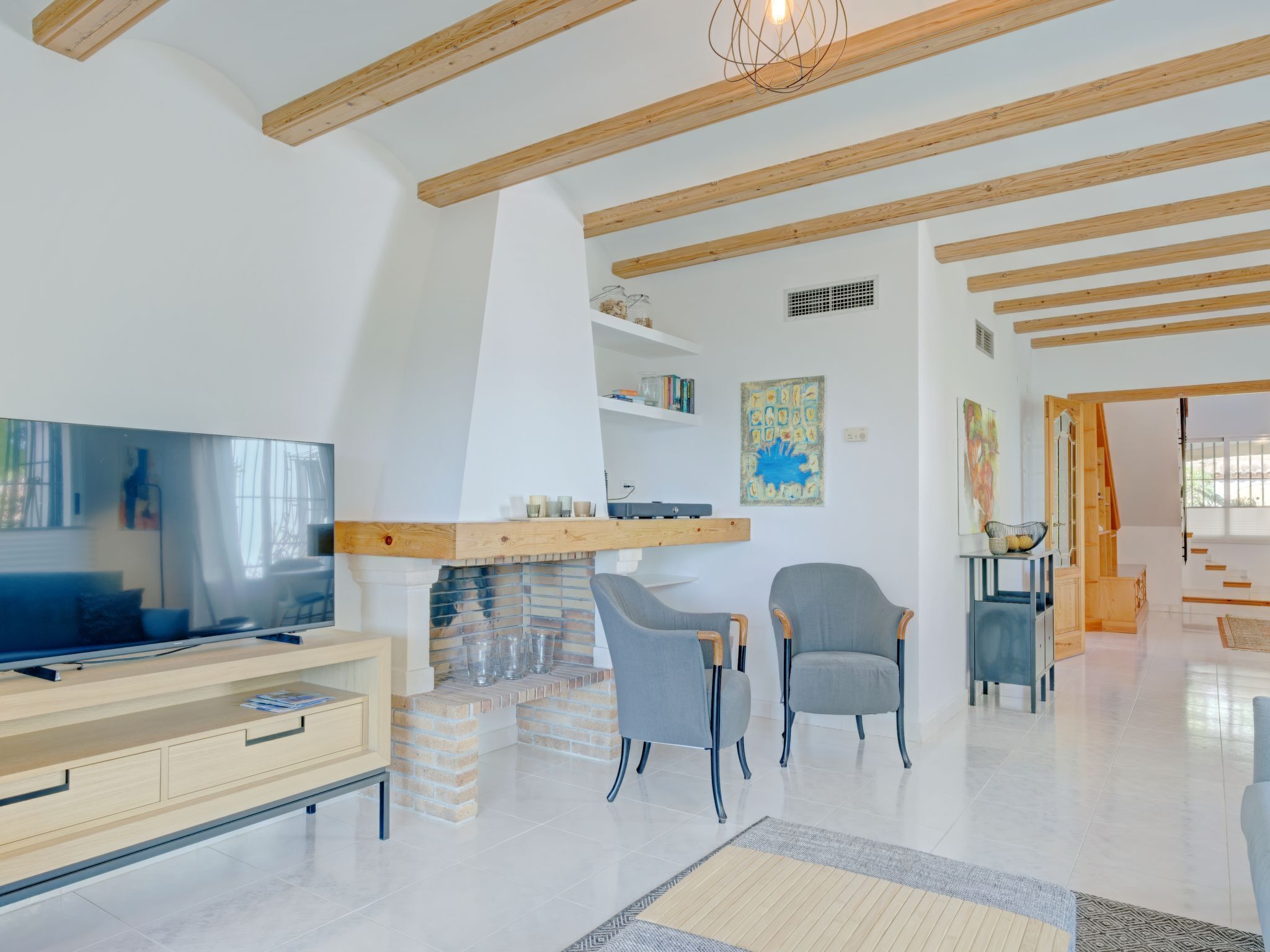 A spacious living room with a TV, two grey chairs, a wooden beam ceiling, and a staircase leading to another level.