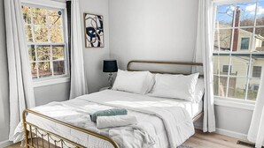 Quarto com cama, mesa de cabeceira, abajur e janela com cortinas.