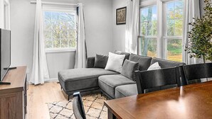 Uma sala de estar moderna com um sofá modular cinza, uma mesa de jantar de madeira e uma grande janela com cortinas.