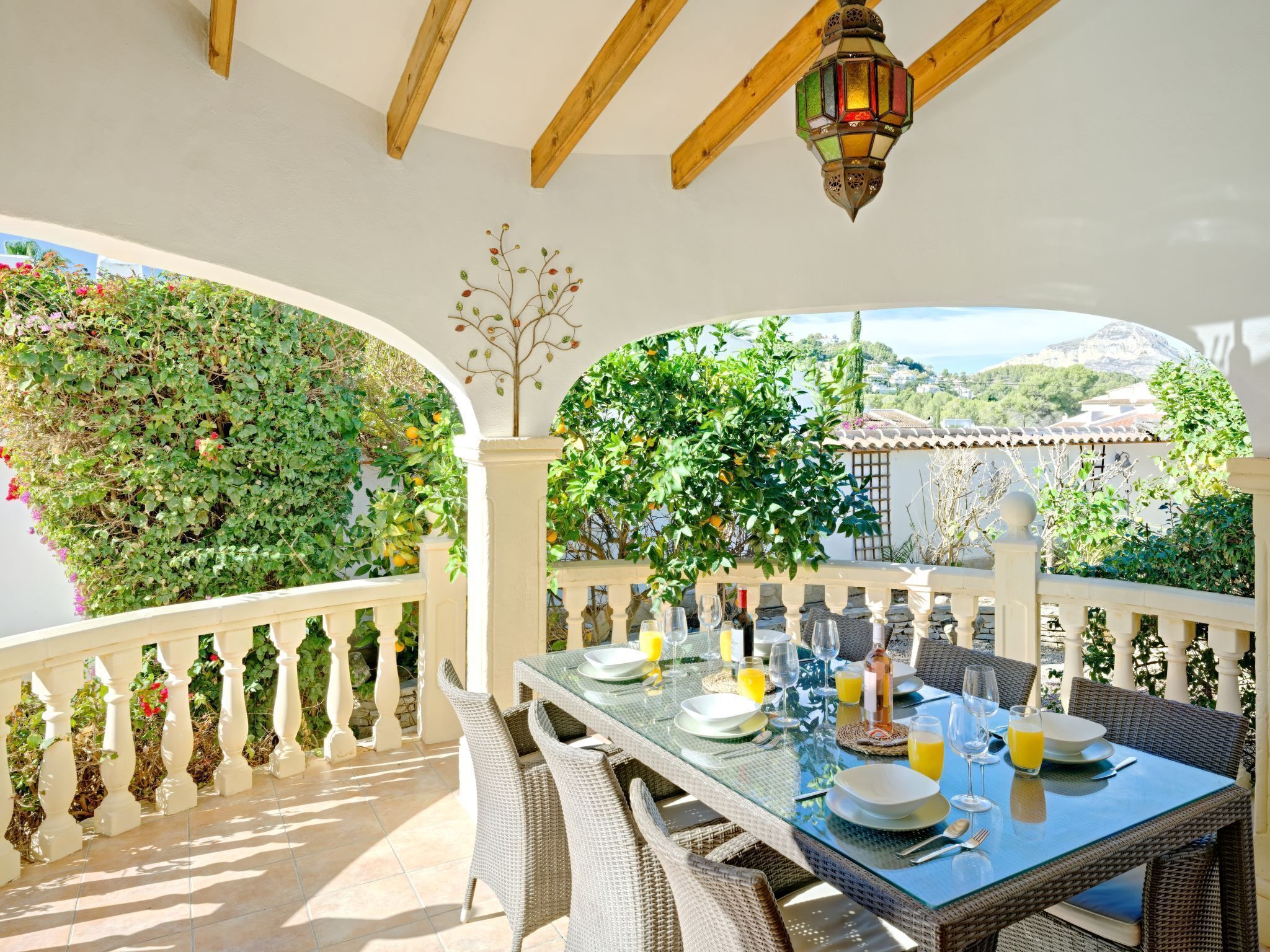 An outdoor dining area with a table set for four, surrounded by wicker chairs, under a pergola with a wooden ceiling and a decorative lantern.