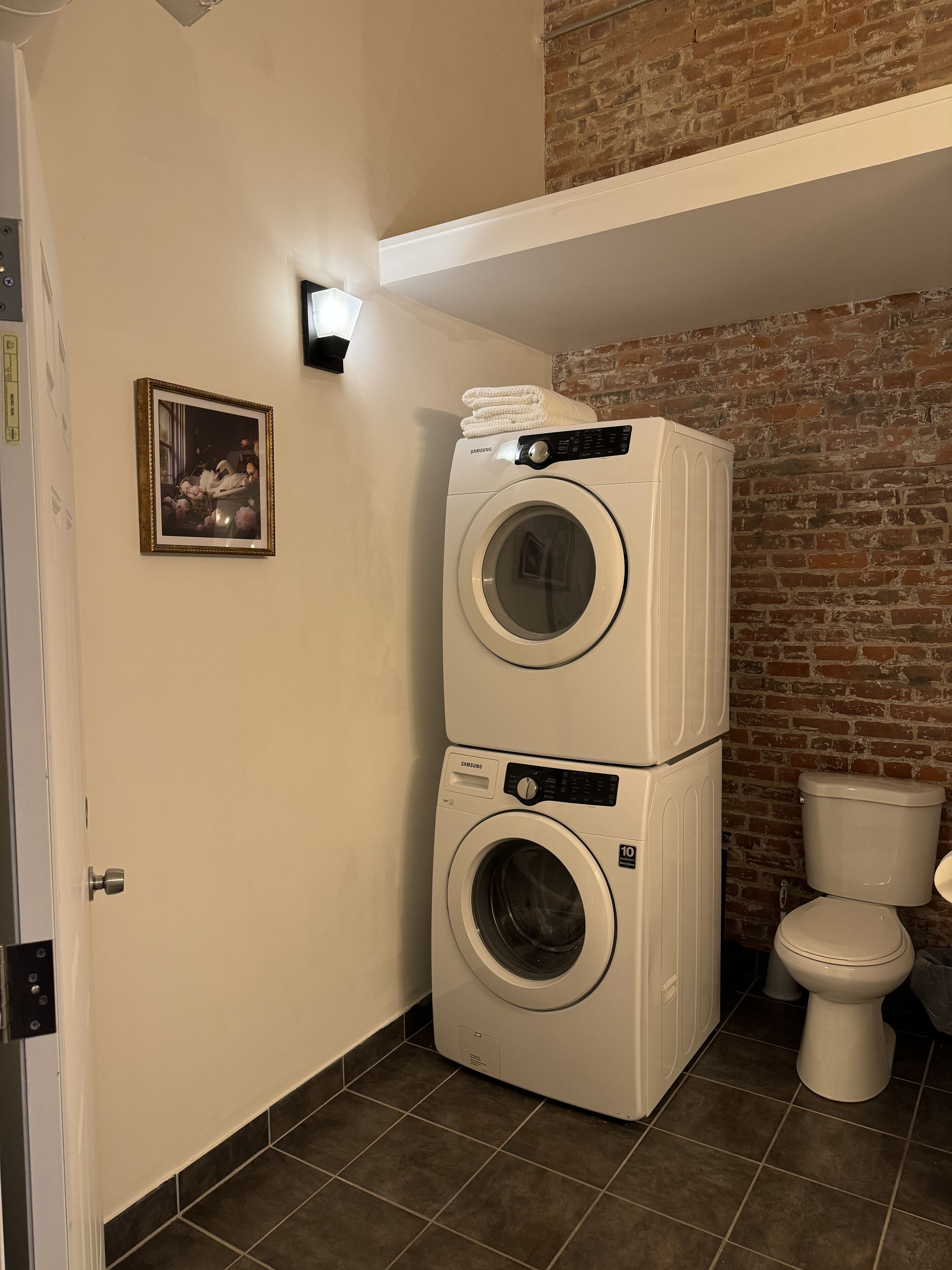Une buanderie avec deux machines à laver et deux sèche-linge blancs superposés, des toilettes, un tableau encadré accroché au mur et un mur en briques.