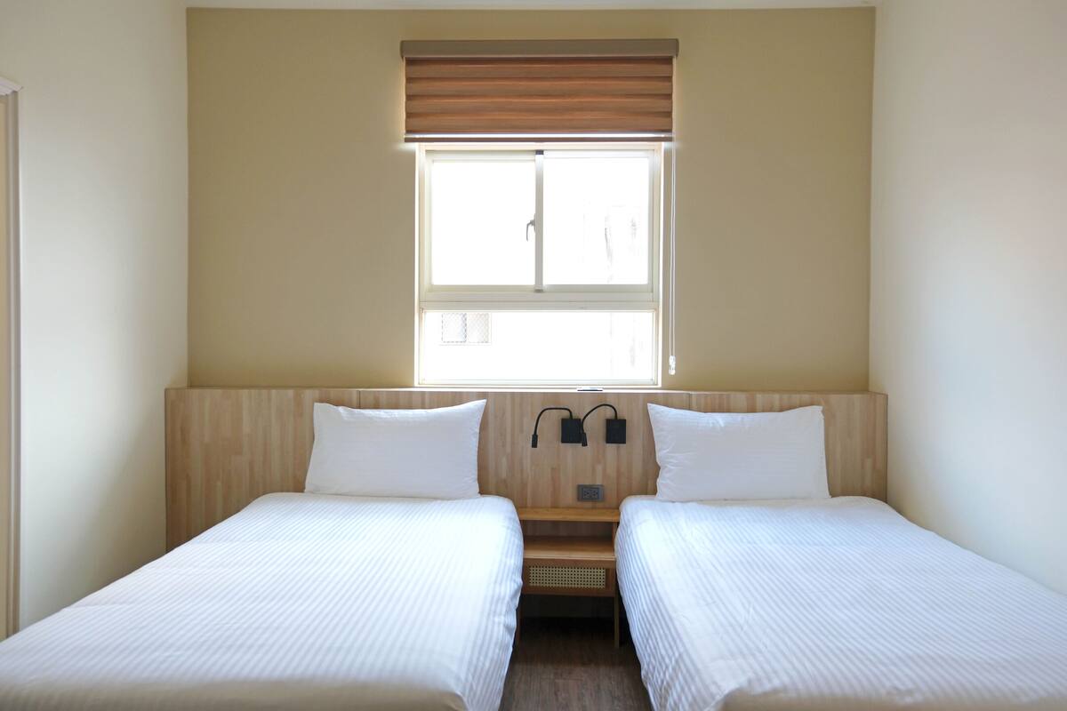 A room with two single beds, a window with blinds, a wooden headboard, and a small shelf.