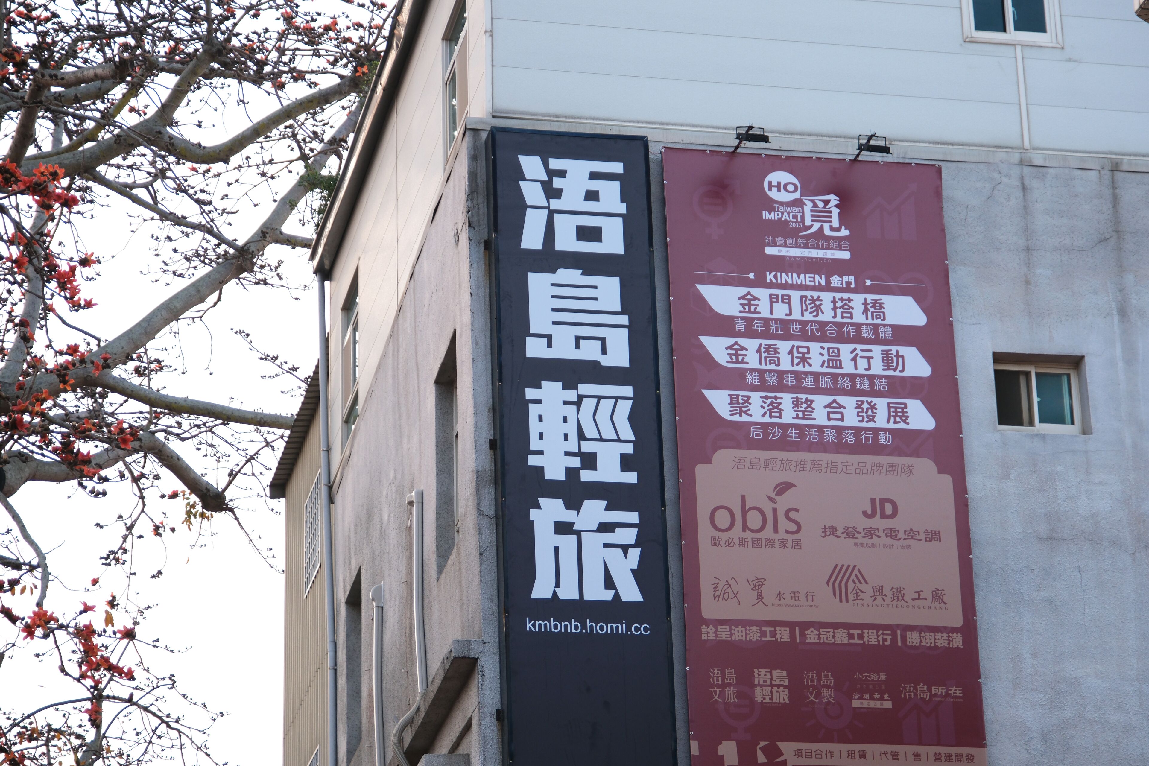 Un edificio con dos pancartas verticales. Una pancarta es negra con caracteres chinos blancos y una dirección web. La otra es roja con caracteres chinos blancos y logotipos.