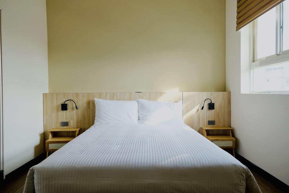 A neatly made bed in a hotel room with a wooden headboard, two bedside tables, and a window providing natural light.