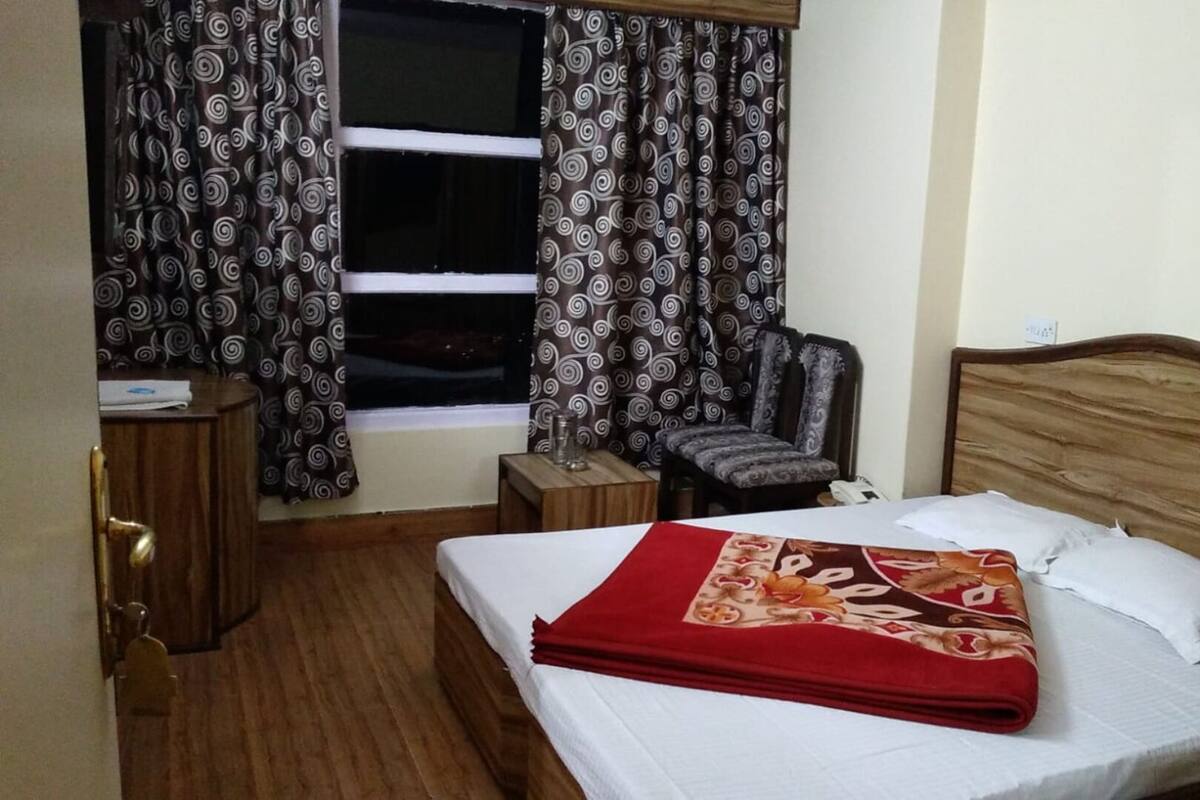 A hotel room with a bed, a chair, a nightstand, a window with curtains, and a wooden floor.