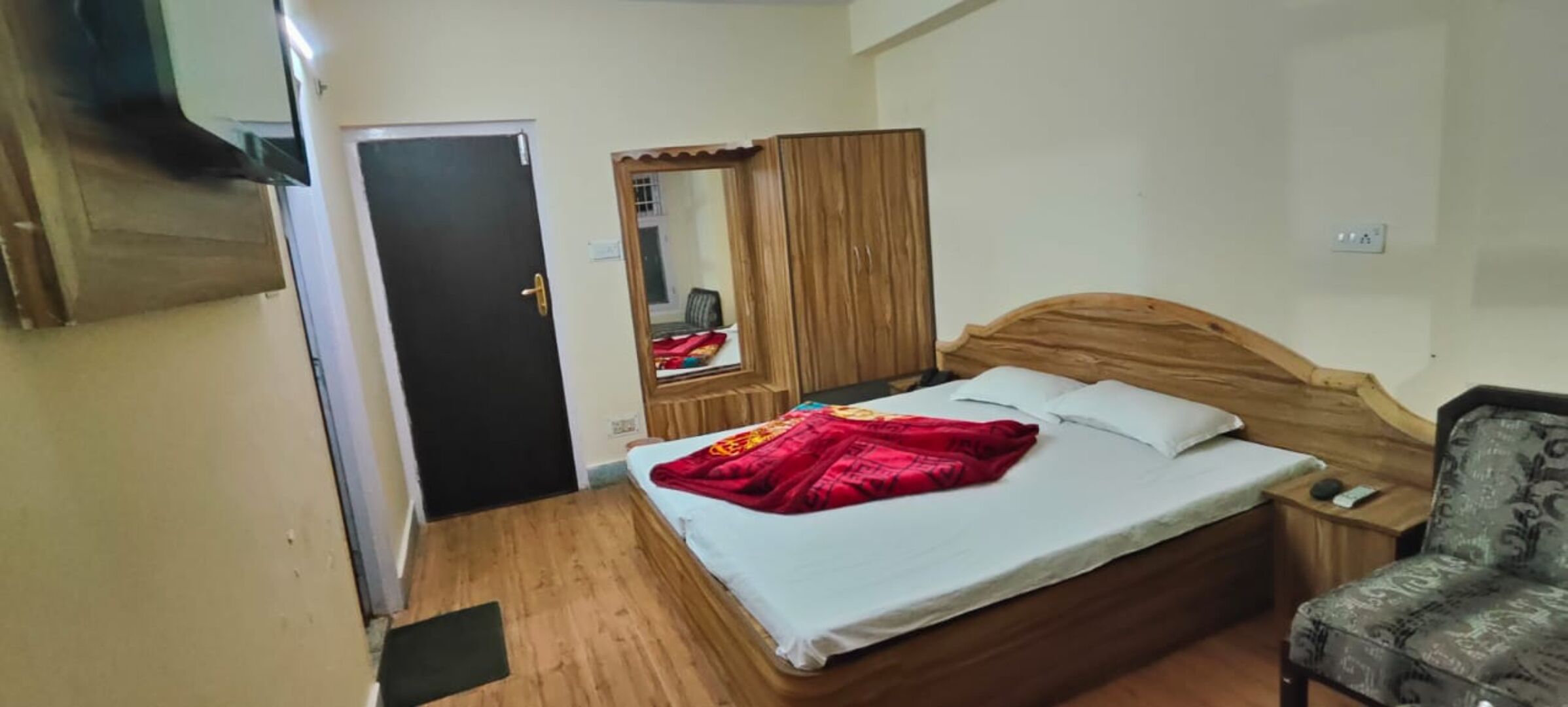 Een slaapkamer met een houten bed, nachtkastje, stoel, kledingkast en een deur.