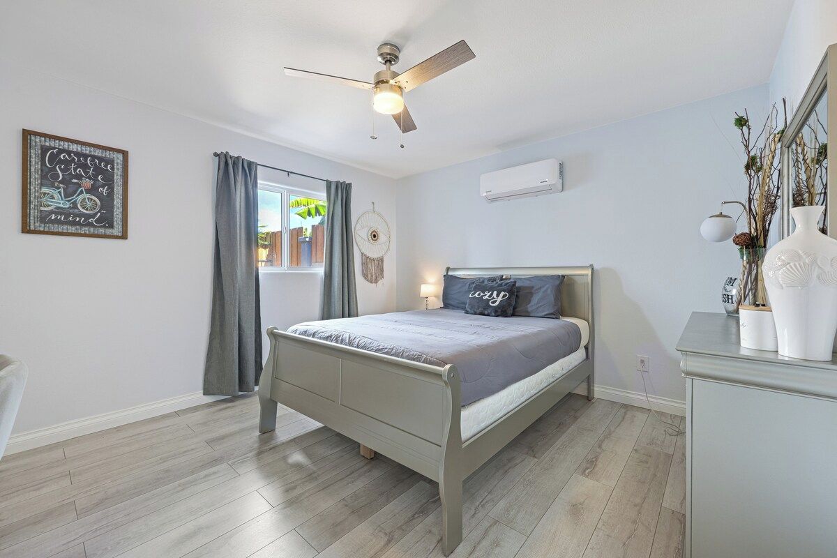 A bedroom with a bed, a ceiling fan, a window with curtains, a wall-mounted air conditioner, and a bedside table with decorative items.