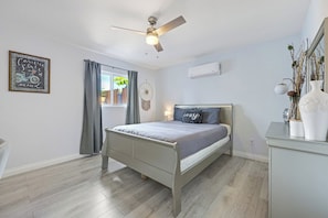 A bedroom with a bed, a ceiling fan, a window with curtains, a wall-mounted air conditioner, and a bedside table with decorative items.