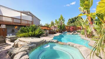 Un patio trasero con piscina, terraza de madera y plantas tropicales.