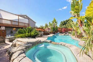 Un patio trasero con piscina, terraza de madera y plantas tropicales.