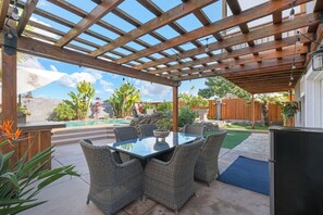 A covered patio with a table and chairs, surrounded by plants and a wooden fence.