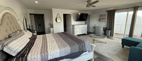 A modern bedroom with a large bed, a TV, a balcony with a view, and a ceiling fan.