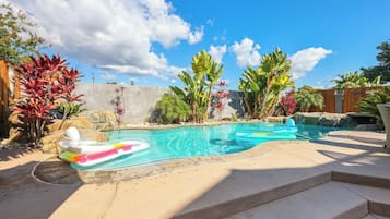 Una piscina con borde de piedra, rodeada de plantas tropicales y una valla de madera.