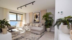 Ett modernt vardagsrum med en beige soffa, ett soffbord och ett stort fönster med utsikt över havet. Det finns två krukväxter och en tavla på väggen.