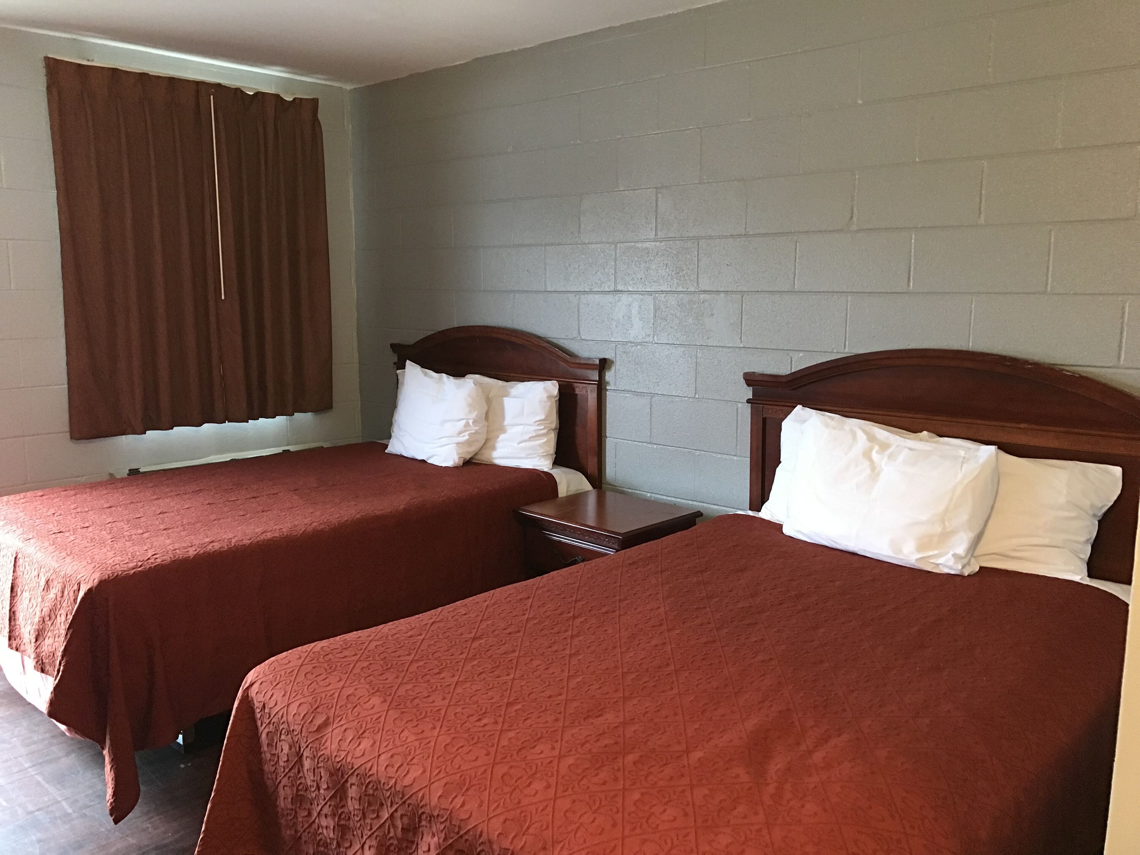 A room with two single beds, each with a wooden headboard and white pillows. The room has a brick wall and a window with brown curtains.
