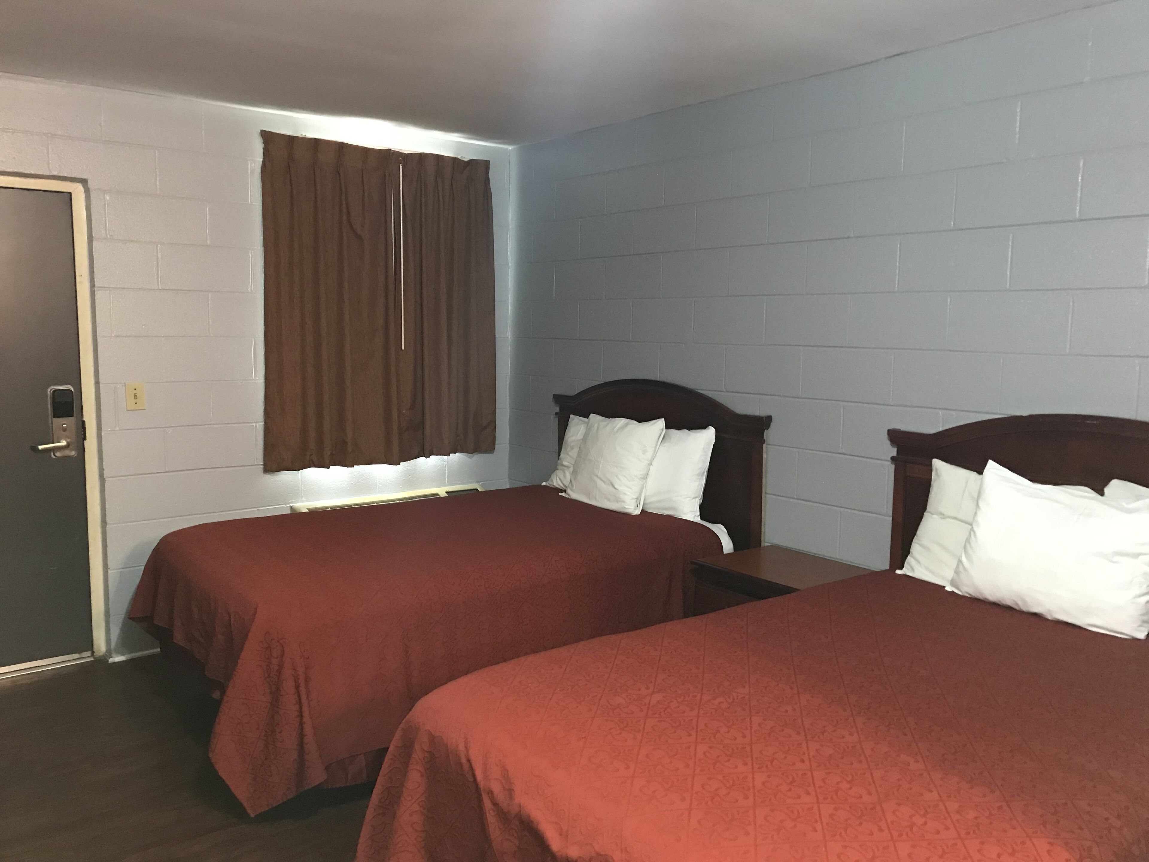 A room with two beds, a window with brown curtains, and a white brick wall.