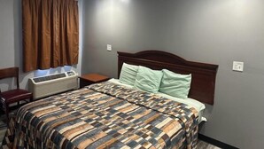 A hotel room with a large bed, a wooden headboard, a window with brown curtains, a bedside table, and a wall-mounted air conditioner. - Sky Inn Hotel (Birmingham)