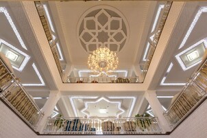 Un atrio de varios niveles con barandillas ornamentadas en blanco y dorado, un candelabro central y diseños geométricos en el techo.