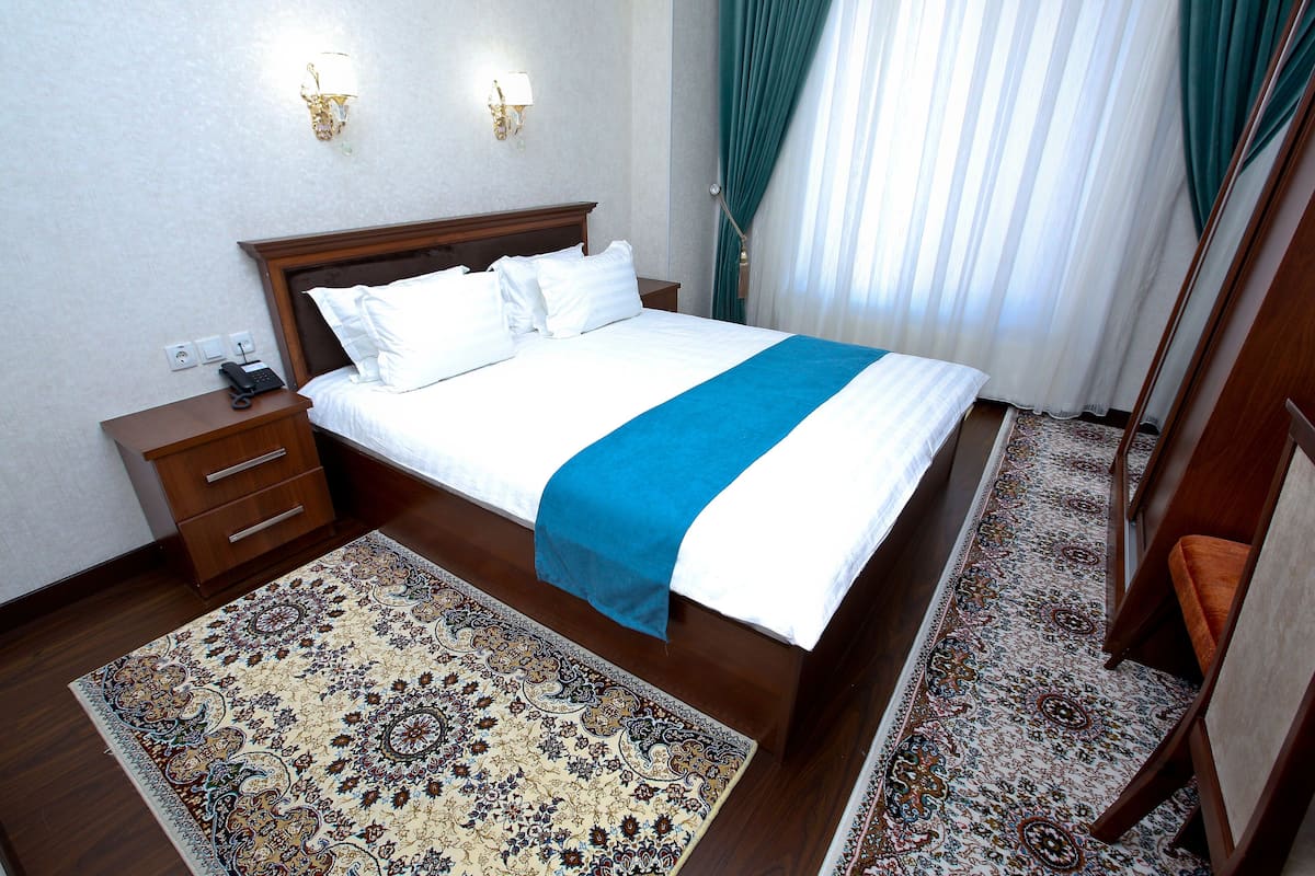 Uma cama bem arrumada com detalhes em azul, uma mesa de cabeceira de madeira com um telefone e um tapete estampado no chão.