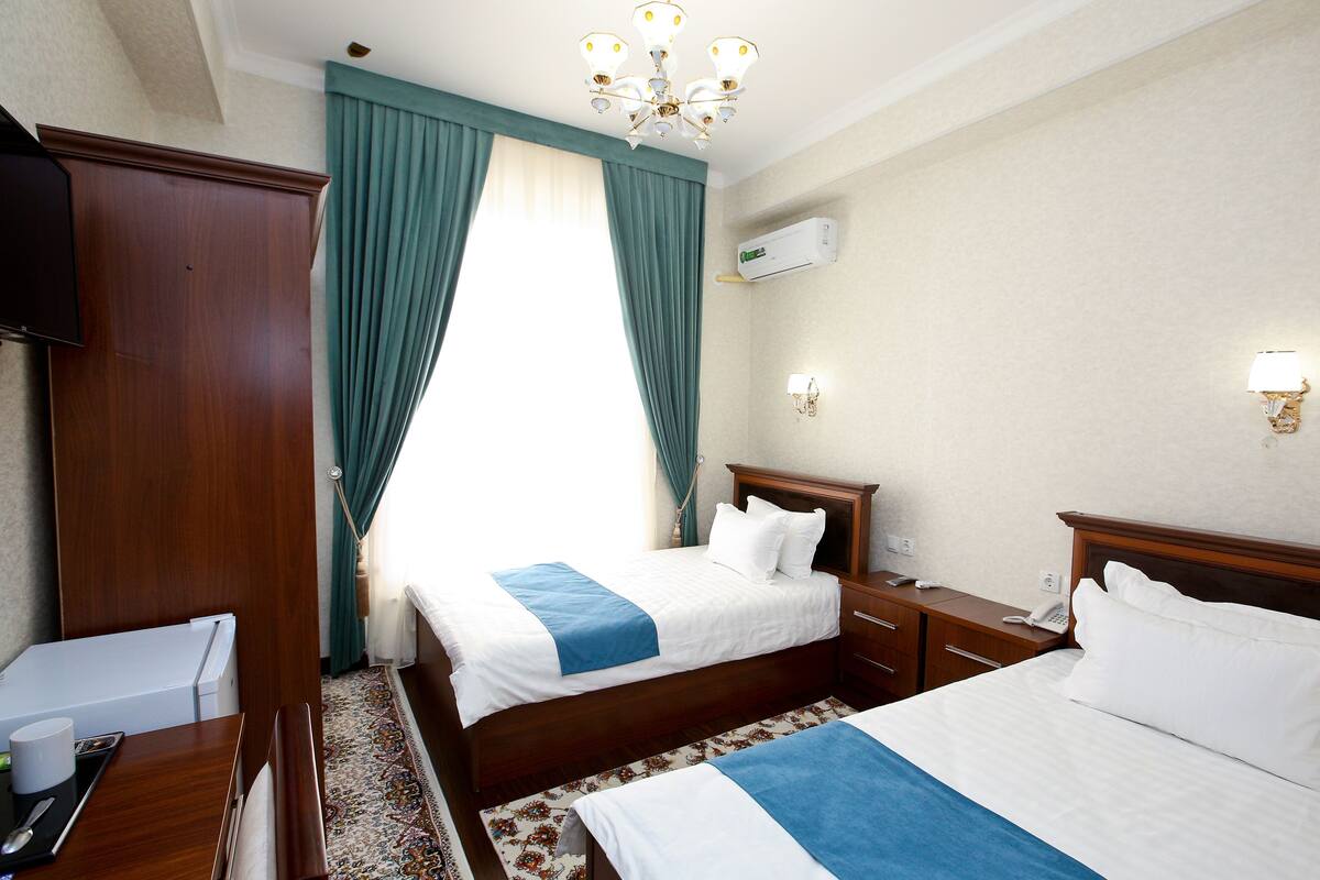 Quarto de hotel com duas camas, uma escrivaninha, um guarda-roupa, um candelabro e uma janela com cortinas.
