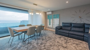A modern living room with a large window overlooking the sea, a dining area with a table and chairs, and a black leather sofa. - SeaLux Apartment - Seaside (Ponta Delgada)