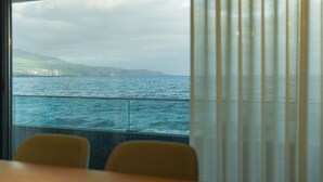 A view from a window with a curtain, overlooking a body of water and distant land with buildings. - SeaLux Apartment - Seaside (Ponta Delgada)