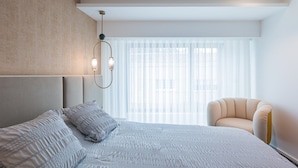 A modern hotel room with a large bed, a sitting area with a chair, and a large window with sheer curtains. - SeaLux Apartment - Seaside (Ponta Delgada)