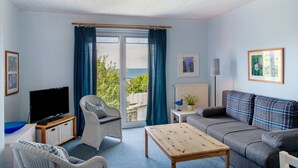 Een woonkamer met een televisie, rieten stoelen, een bank, een tafel en uitzicht door een groot raam.