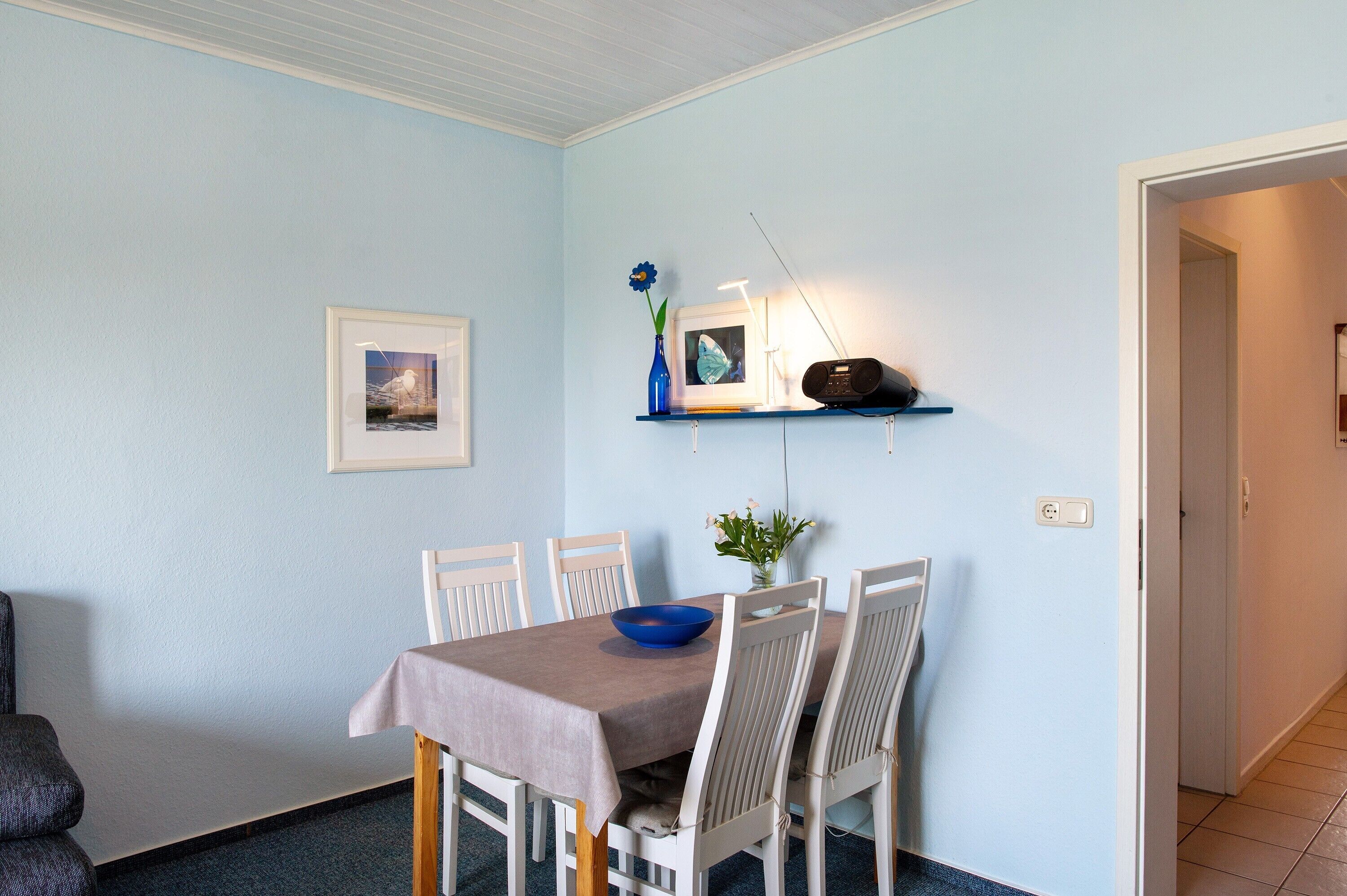 Ein Essbereich mit Tisch, Stühlen, einer Blumenvase, einem gerahmten Bild und einem Regal mit einem Radio.