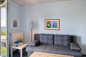Una habitación con un sofá gris, una pequeña mesa con una lámpara, un cuadro enmarcado y una planta en maceta.