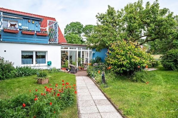 A house with a red roof, a blue exterior, and a garden with red flowers and a pathway leading to the entrance.