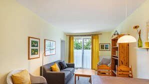 Una sala de estar con sofá, sillón, mesa de comedor y sillas. Hay una estantería y una ventana con cortinas.