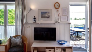 Un salon avec un téléviseur, un fauteuil en osier, un bol bleu et un tableau encadré au mur.