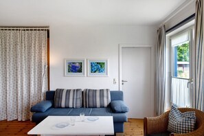 Quarto com sofá azul, mesa de centro branca e cadeira de vime. Há duas fotos emolduradas na parede, uma porta e uma janela com cortinas.